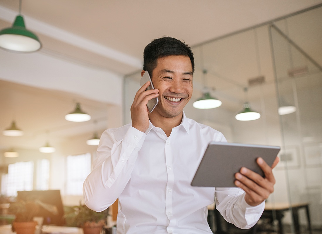 Business Insurance - Portrait of a Smiling Young Businessman Standing in a Conference Room in a Modern Office While Talking on the Phone and Holding a Tablet